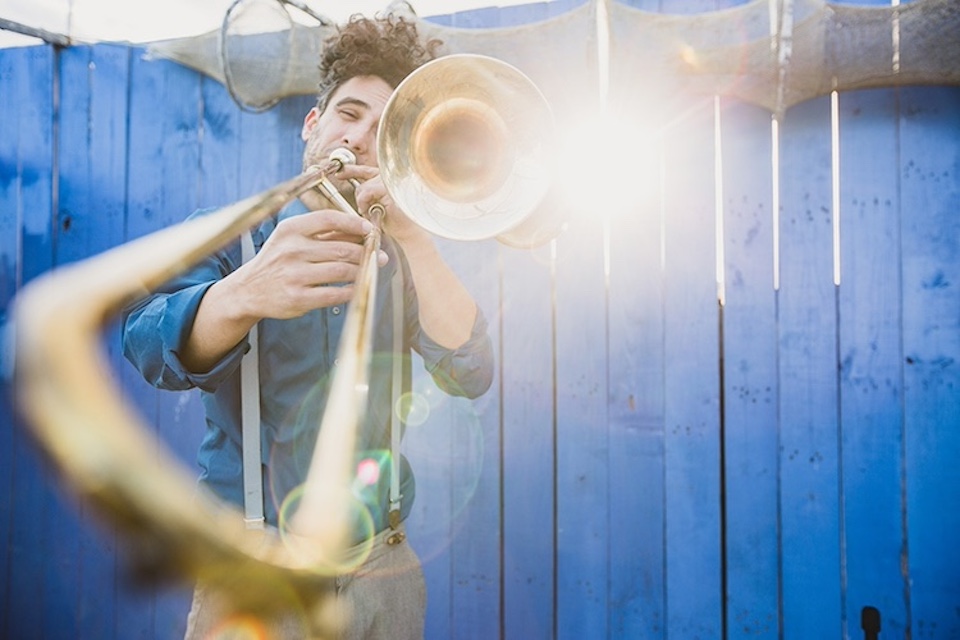 Eric Tarantola spielt ein Blasinstrument vor einem blauen Zaun.
