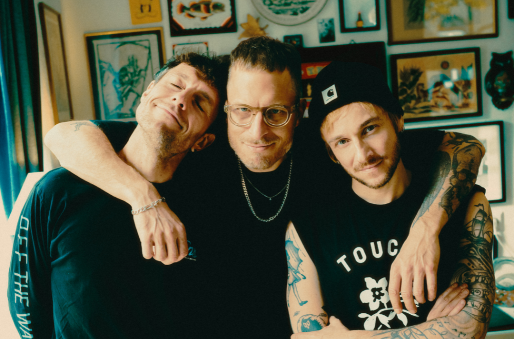 Die drei Musiker der Band ITCHY stehen vor einer Bilderwand.