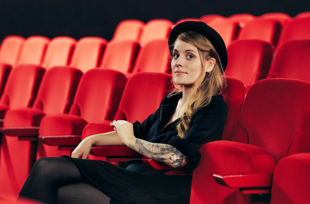 Laura Braun sitzt in einem Theatersaal, um sie sieht man leere, rote Theatersessel