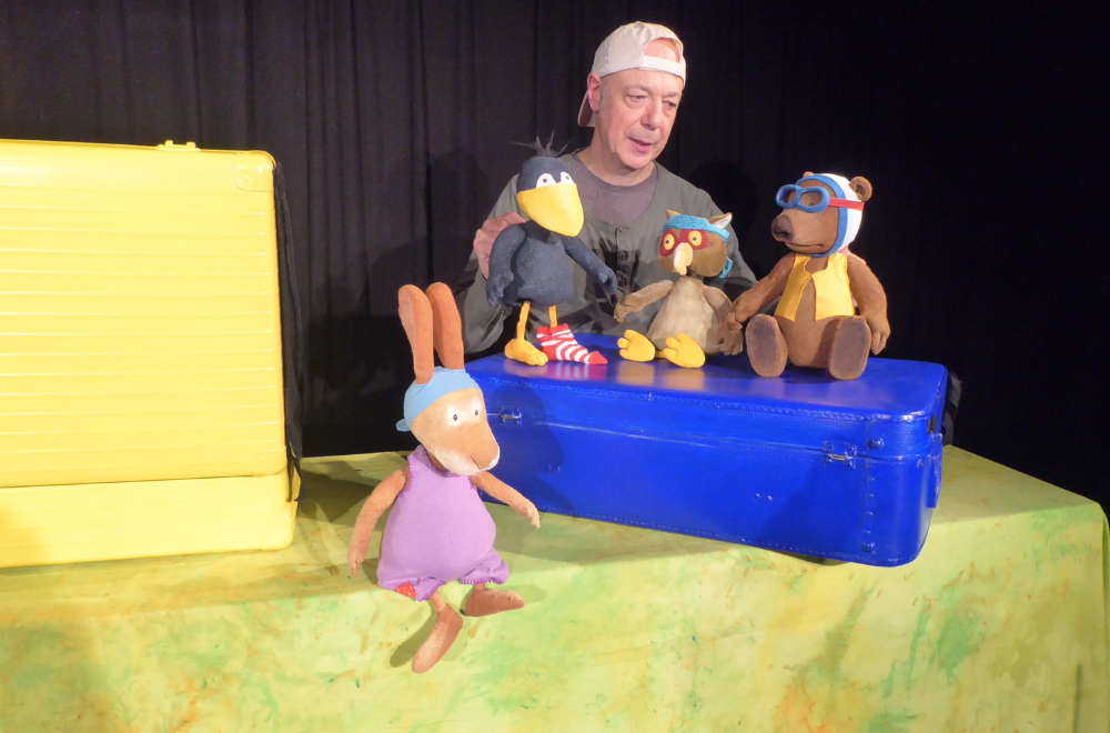 Der kleine Rabe Socke und andere Puppenfiguren sitzen auf einem blauen Koffer.